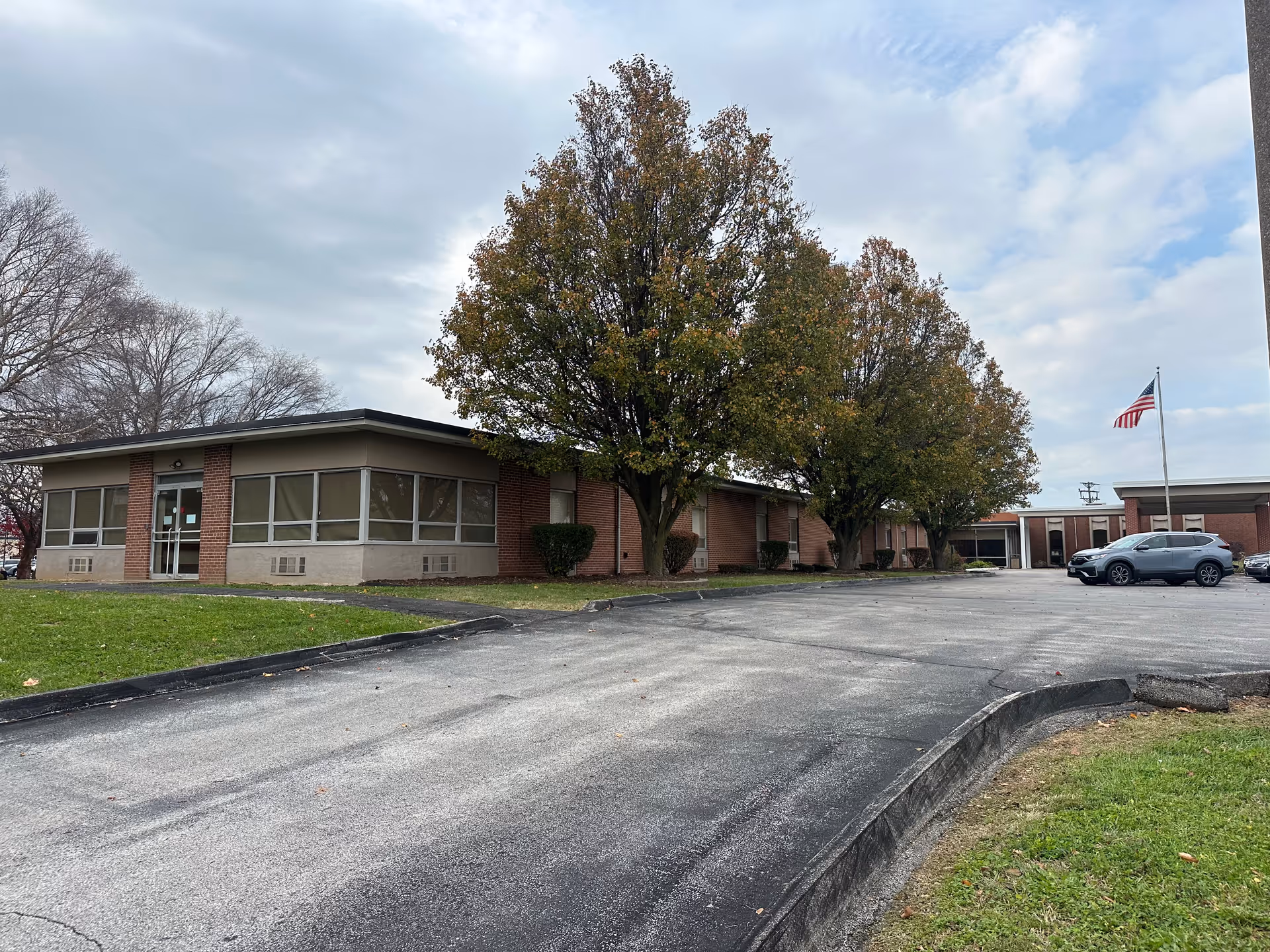 Exterior view of a single-story brick building with large windows and a driveway leading up to the entrance. Several trees with autumn foliage are in front of the building, and an American flag is flying on a flagpole near the parking area with a few parked cars.