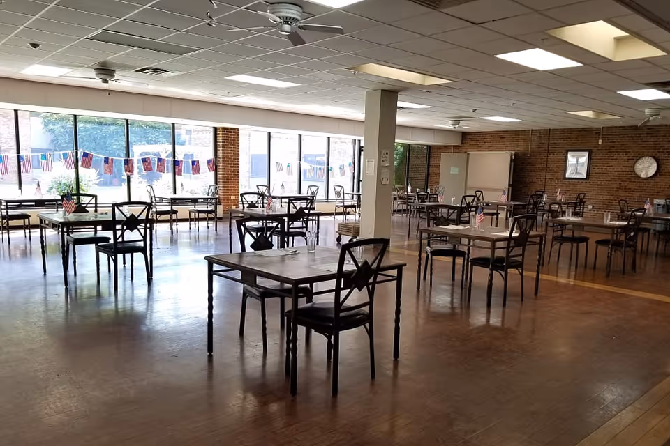 A spacious dining room with multiple tables and chairs arranged neatly. Each table has a small American flag and a glass of water. Large windows along one wall let in natural light, and patriotic decorations hang across the windows. The room has a brick wall with a clock and framed picture, and ceiling fans are visible.