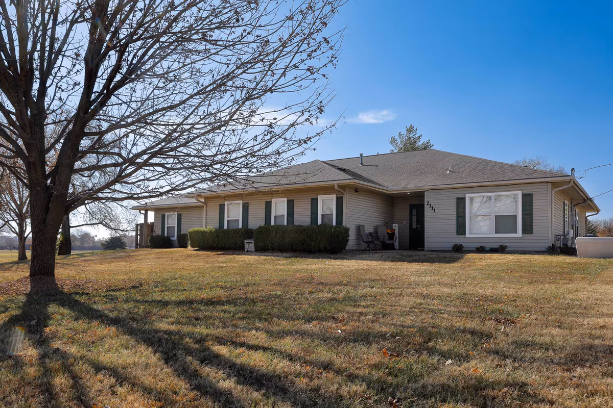 Single-story beige ranch-style building with green shutters on a grassy yard and a large leafless tree in the foreground.