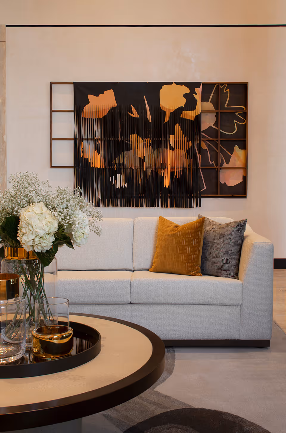 Modern living room with a white sofa, decorative pillows, a round coffee table with glassware and flowers, and abstract wall art on the wall.