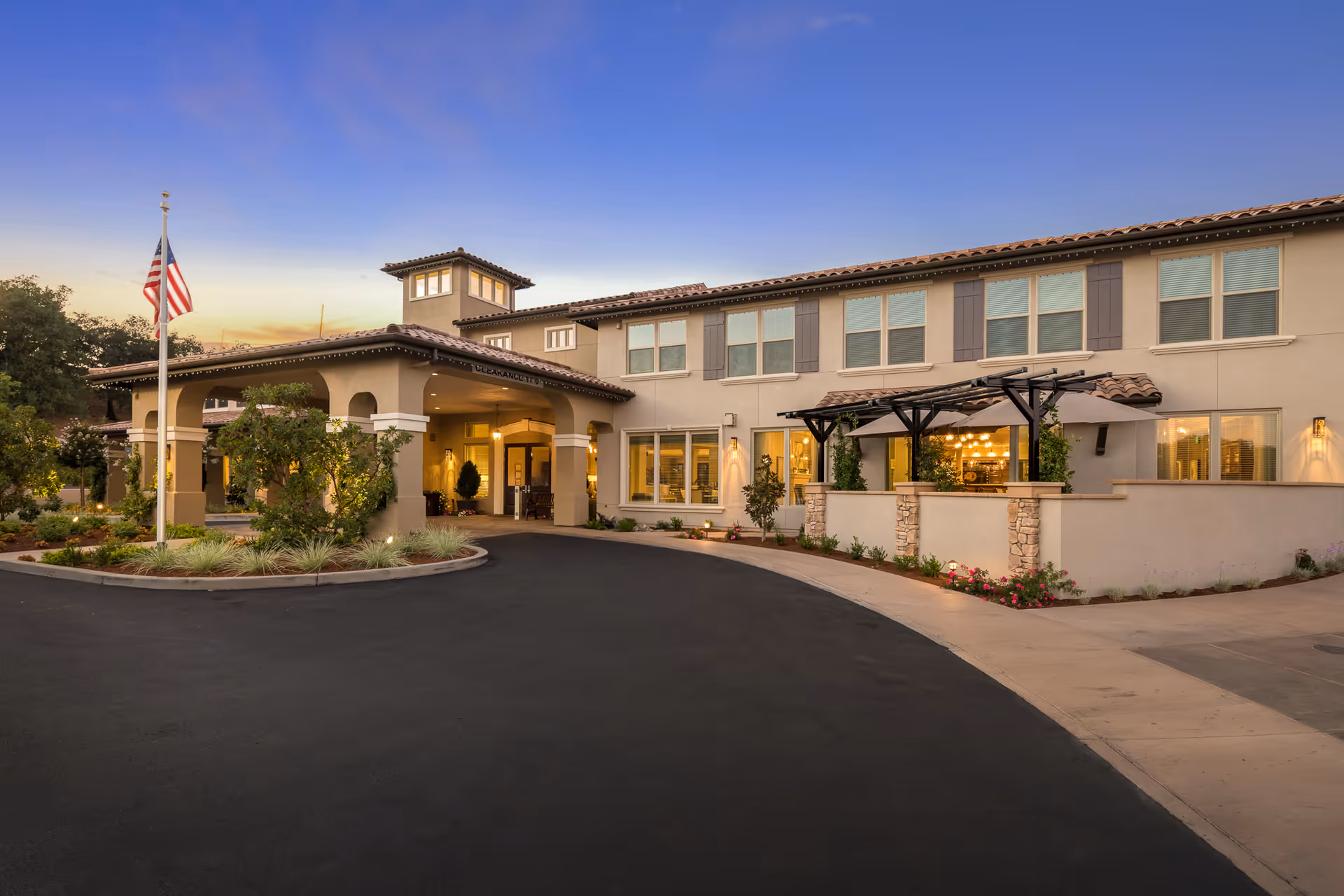 Exterior view of Villa Capri at Varenna senior living facility during sunset, showing a two-story building with a covered entrance, landscaped garden, American flag, and warm lighting inside.
