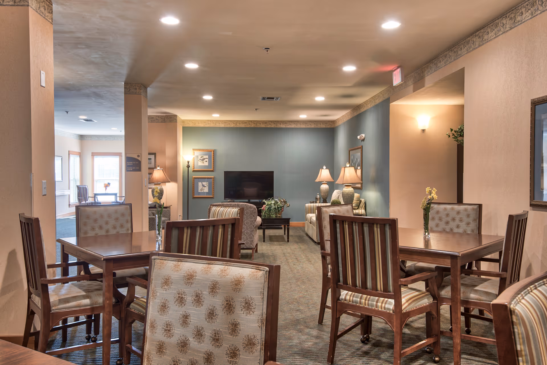 Interior view of a senior living facility common area with several wooden tables and chairs arranged for seating. The room has soft lighting with lamps and ceiling lights, a television mounted on a blue accent wall, framed artwork, and a carpeted floor. The space appears cozy and inviting with neutral and warm tones.