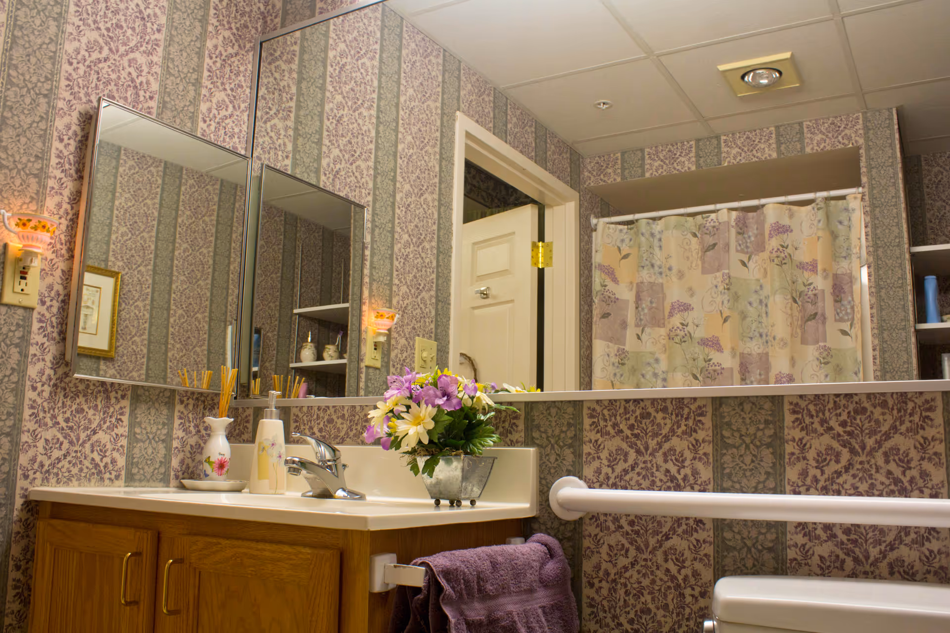 Bathroom interior with floral and striped wallpaper, a wooden vanity with a sink, a faucet, and decorative items including a vase with flowers and a soap dispenser. There are two mirrors on the wall, a towel hanging on a white grab bar, and a shower curtain with a floral pattern in the background.