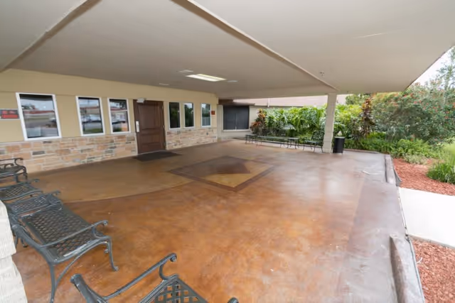 Covered entrance/porte-cochere with metal benches leading to a wooden door and landscaping at the side.