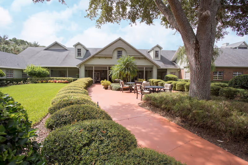 Front entrance courtyard with a paved walkway, trimmed hedges, outdoor seating, and a low building with dormer windows.
