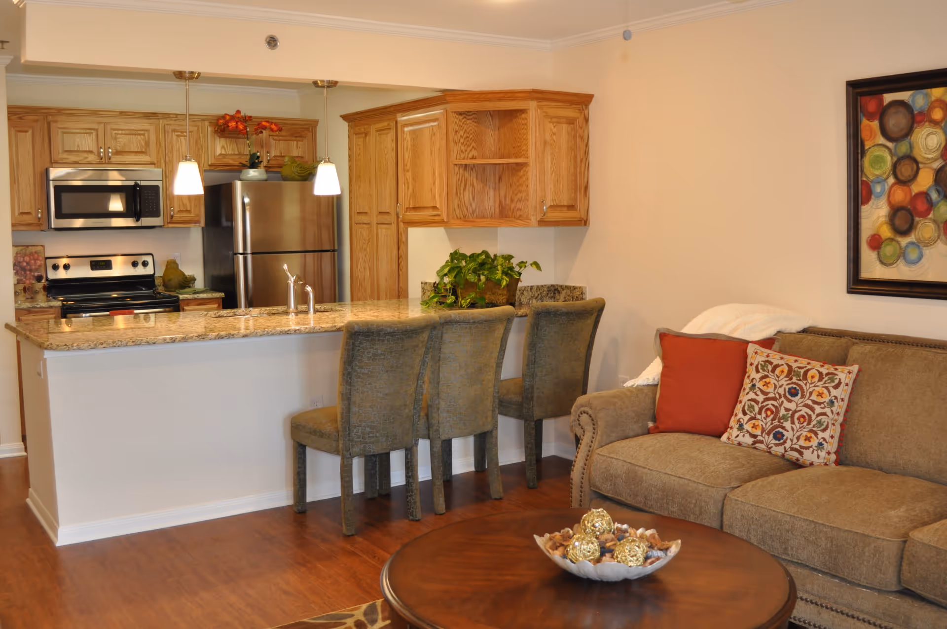 A cozy living area with a brown sofa adorned with red and patterned cushions, a round wooden coffee table with decorative items, and a kitchen area with granite countertops, wooden cabinets, stainless steel appliances, and three upholstered bar stools.