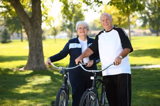 An elderly man and woman standing outdoors in a park-like setting, each holding a bicycle and smiling at the camera with trees and grass in the background.
