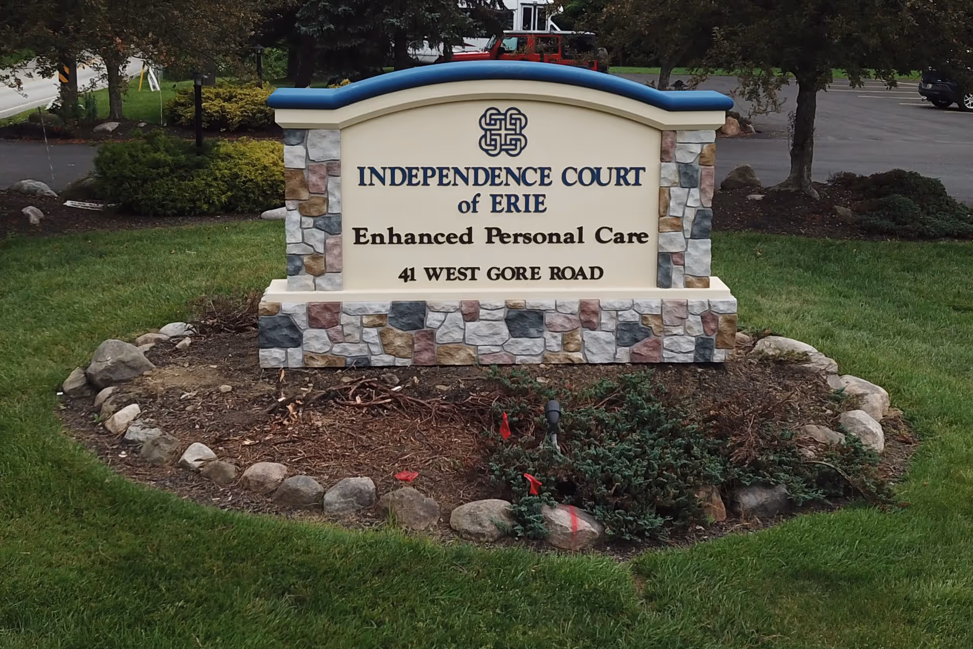 Stone and brick sign for Independence Court of Erie Enhanced Personal Care located at 41 West Gore Road, surrounded by grass and landscaping with a parking lot and trees in the background.