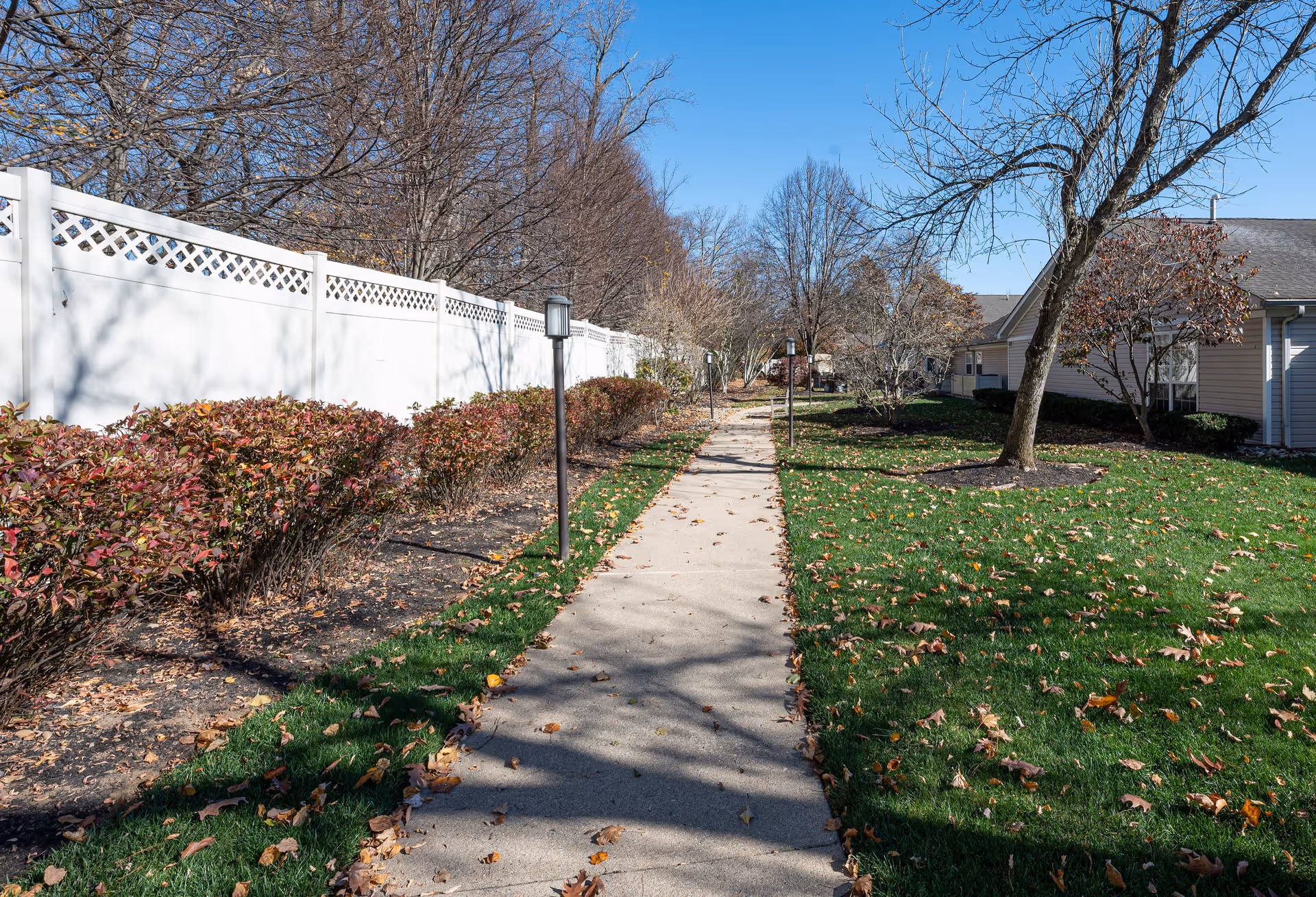 A paved walkway lined with small lamp posts runs through a landscaped outdoor area with green grass, bushes, and leafless trees. A white fence is on the left side, and residential-style buildings are on the right under a clear blue sky.