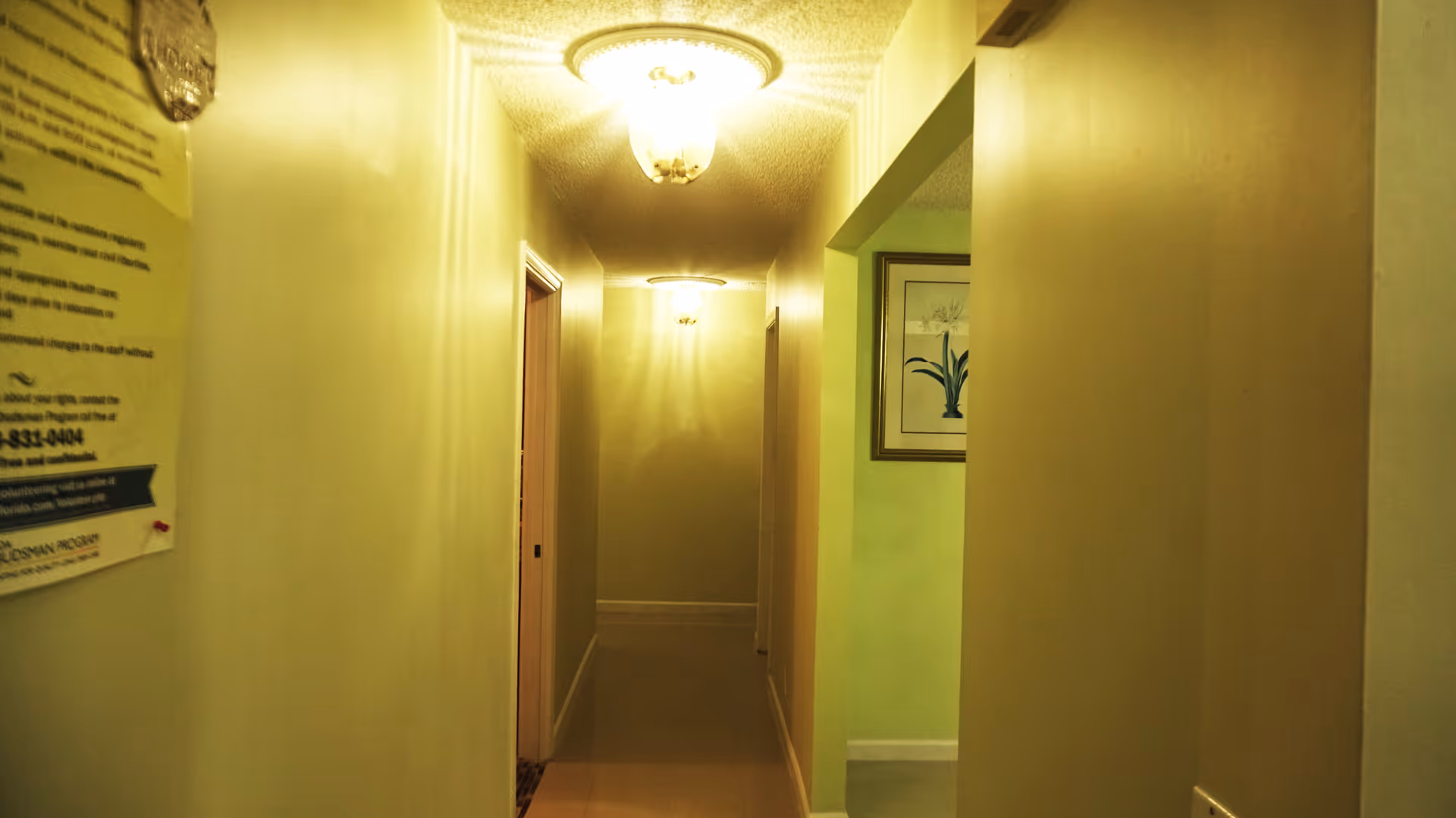 A narrow hallway in an assisted living facility with beige walls and ceiling lights. There are doorways on both sides of the hallway and a framed picture on the right wall. A notice is partially visible on the left wall near the entrance of the hallway.