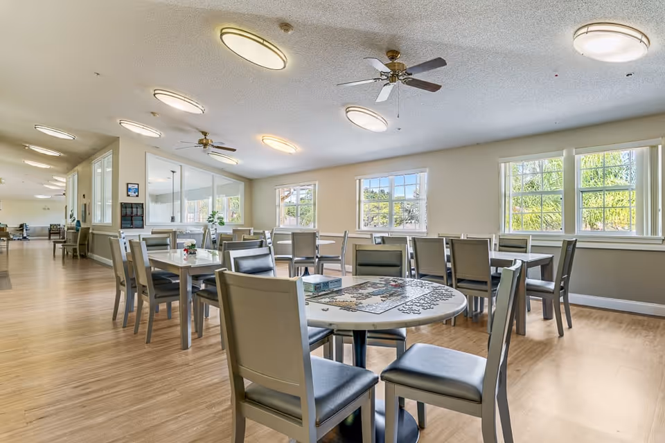 A bright and spacious common area in a senior living facility with multiple tables and chairs arranged for seating. One round table in the foreground has a partially completed jigsaw puzzle on it. The room has large windows letting in natural light, ceiling fans, and overhead lights. The floor is wooden, and the walls are painted in neutral tones.