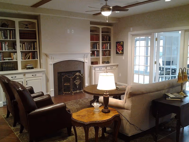 A cozy living room with two dark brown leather armchairs, a beige sofa, a wooden coffee table with a lamp, built-in bookshelves filled with books, a fireplace with a decorative screen, and glass double doors leading to another room.