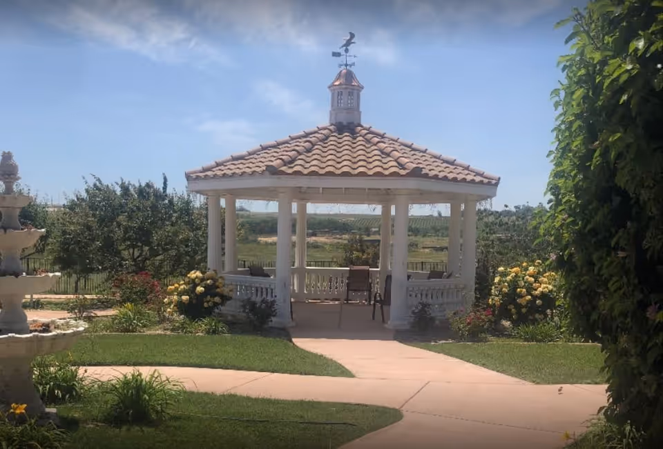 A white gazebo with a tiled roof and a weather vane on top, situated in a garden with green grass, flowering bushes, and a paved walkway leading to it. There are chairs inside the gazebo and a multi-tiered water fountain visible on the left side. The sky is clear and blue.