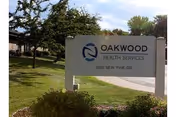 Outdoor view of a sign for Oakwood Health Services located at 2912 New Pine Dr, with trees, grass, and a building partially visible in the background under a partly cloudy sky.