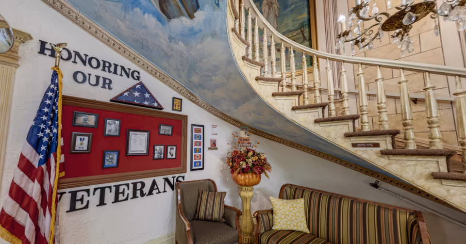 Interior view of a senior living facility showing a curved staircase with ornate railing and a painted ceiling. Below the staircase is a seating area with a striped couch and an armchair. On the wall, there is a display honoring veterans with framed photos, medals, and an American flag. A floral arrangement is placed on a pedestal next to the seating area.