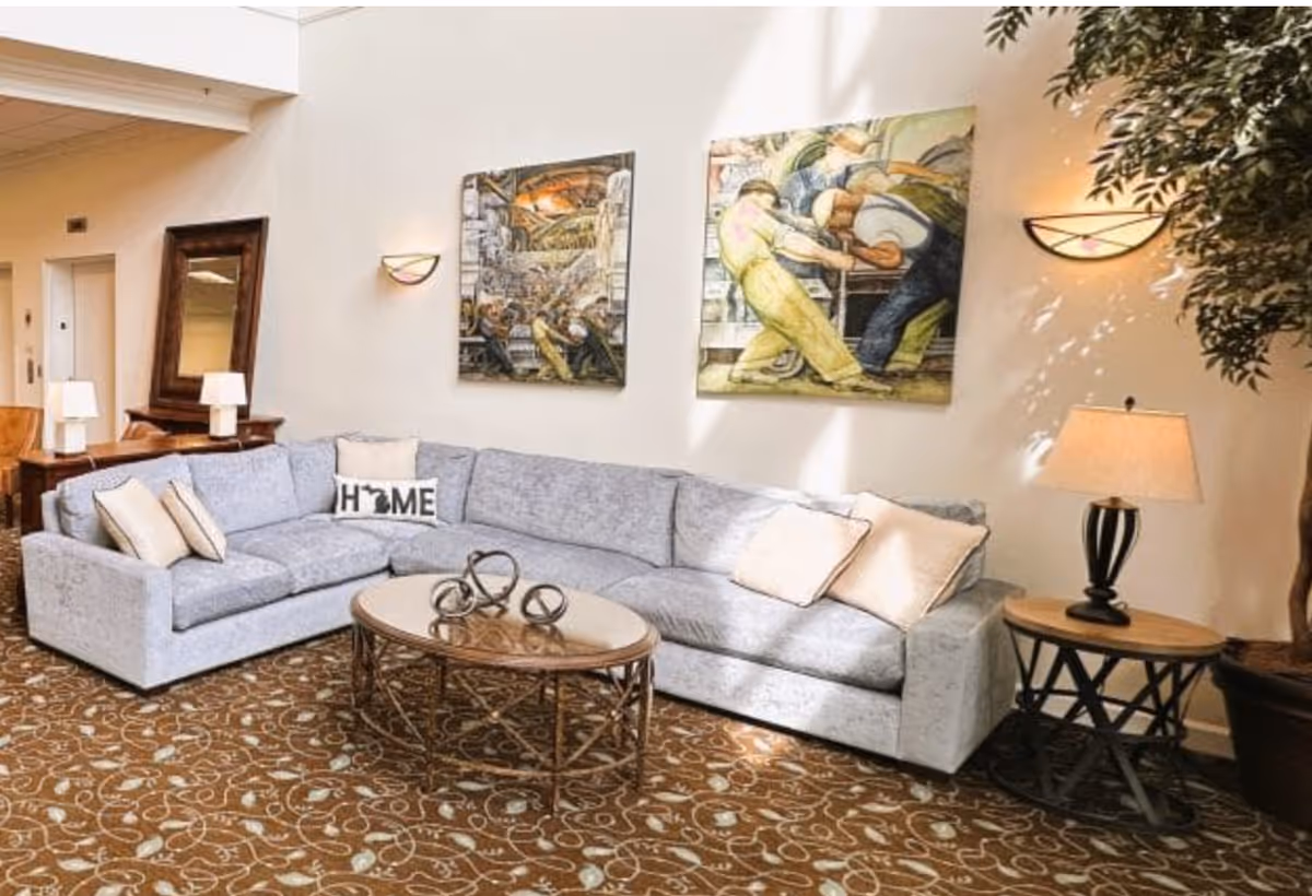 A cozy living room area with a large light gray sectional sofa adorned with several pillows, including one with the word 'HOME' and a silhouette of Michigan. A round wooden coffee table with decorative metal rings sits in front of the sofa. The walls are decorated with two colorful paintings depicting people working. There are two wall-mounted lights and a table lamp on a round side table next to the sofa. A large mirror and additional lamps are visible in the background, along with a tall potted plant on the right side.