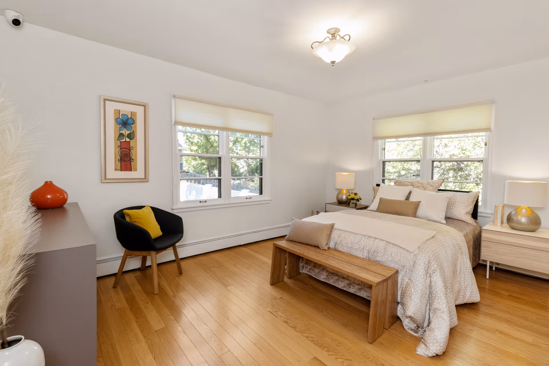 A bright and cozy bedroom with a large bed covered in beige and white bedding, two bedside tables with lamps, a wooden bench at the foot of the bed, a black chair with a yellow cushion, a dresser with a red vase, and two windows with white blinds letting in natural light.