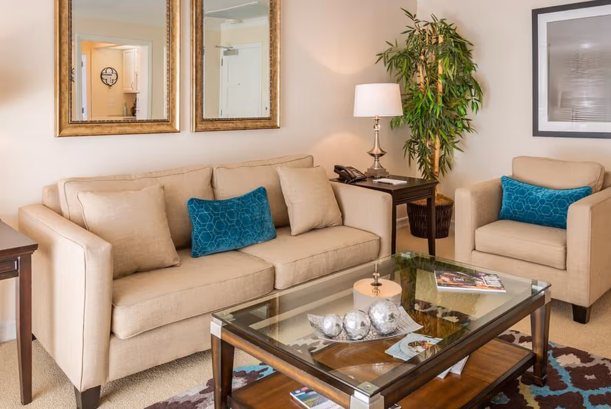 A cozy living room with a beige sofa and matching armchair, both adorned with teal accent pillows. A glass coffee table with decorative silver spheres and magazines sits in front of the sofa. Two large gold-framed mirrors hang on the wall above the sofa. A side table with a lamp and a telephone is placed next to the armchair, and a tall green plant is in the corner. A framed black and white picture is on the wall to the right.
