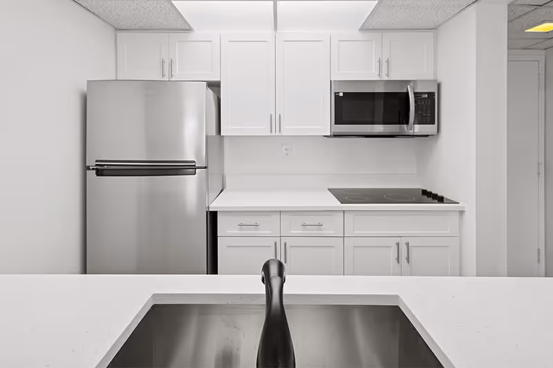 Modern kitchen with white cabinets, a stainless steel refrigerator, a built-in microwave, an electric stovetop, and a sink with a black faucet in the foreground.