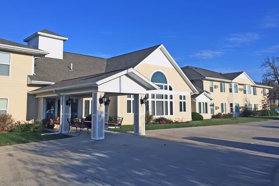 Exterior view of a light-yellow assisted living building with a covered entrance, benches, and a paved driveway under a clear blue sky.