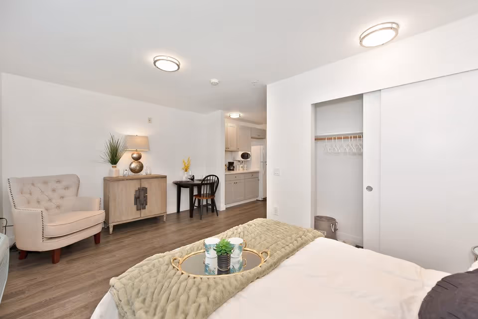 A bright and clean senior living studio apartment featuring a bed with a green throw blanket and a tray with cups and a small plant. There is an open closet with hangers and a trash bin, a small wooden cabinet with a lamp and plant on top, a beige armchair, and a small black table with a chair. The kitchenette with white cabinets and appliances is visible in the background. The room has wood flooring and white walls with ceiling lights.