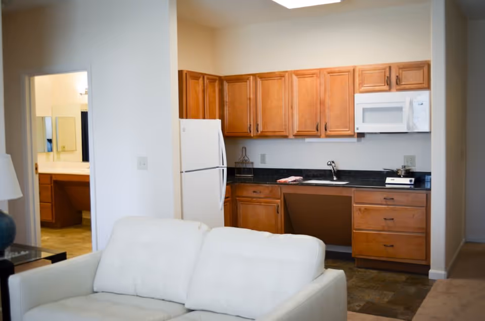 Interior view of a living space featuring a white couch in the foreground, a kitchen area with wooden cabinets, a white refrigerator, microwave, and a sink in the background. To the left, there is an open doorway leading to a bathroom with a vanity and mirror visible.