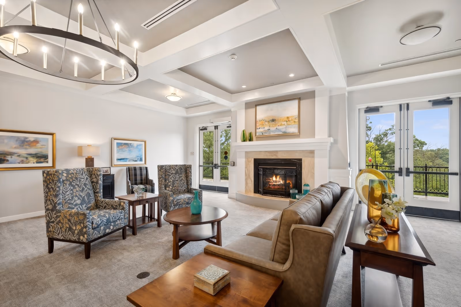 A bright and spacious living room area with a beige leather sofa, two patterned armchairs, a round wooden coffee table, and a fireplace with a painting above it. Large glass doors provide a view of greenery outside. The room features a modern chandelier and neutral-colored walls and carpet.
