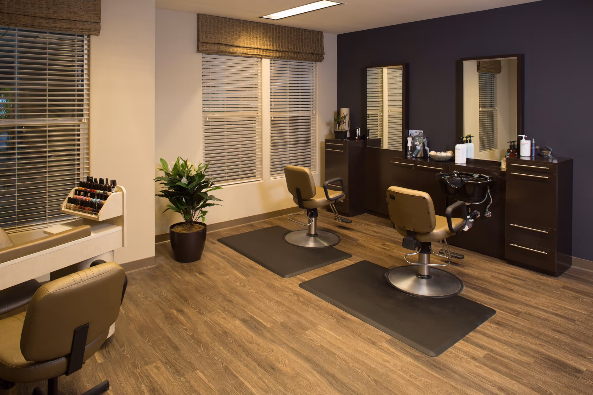 Interior view of a salon area with two styling chairs in front of mirrors and a counter with hair care products. There is a potted plant near the window with blinds and a rack of nail polish on the left side. The floor is wooden with mats under the chairs.