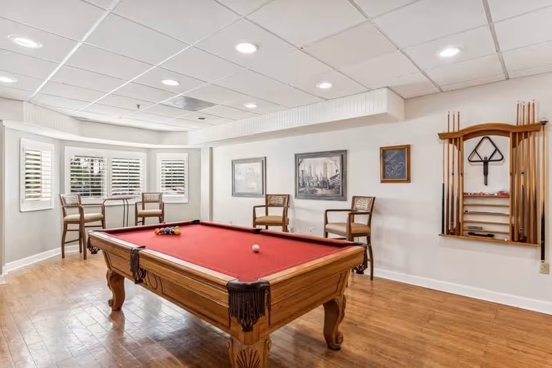 Interior room with a red felt pool table in the center, surrounded by wooden chairs and a small table near windows with white shutters. The walls have framed pictures and a mounted rack holding pool cues and a triangle rack.