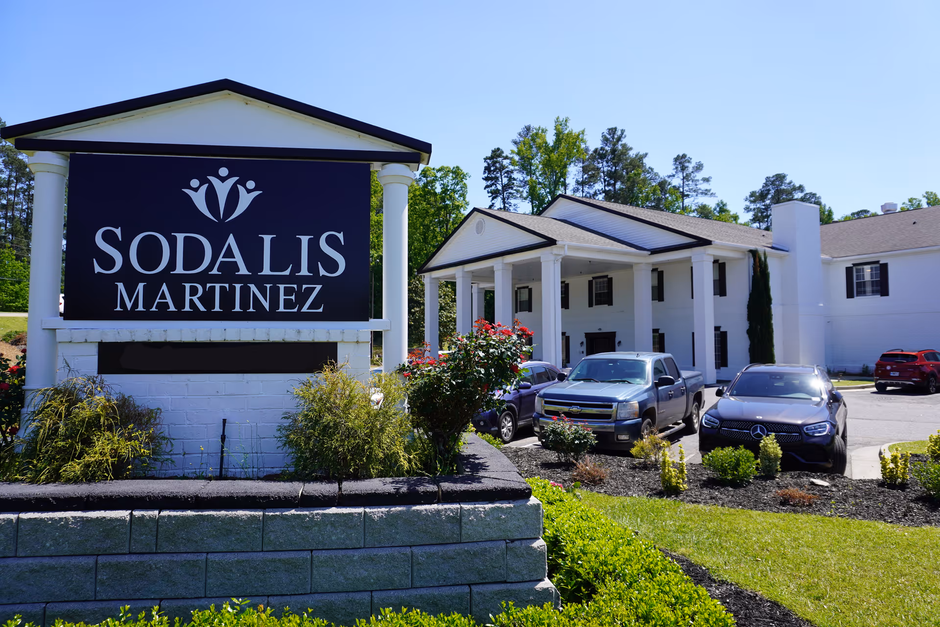 Exterior view of Sodalis Martinez senior living facility showing a large sign with the facility name in front of a white building with columns, surrounded by greenery and parked cars.