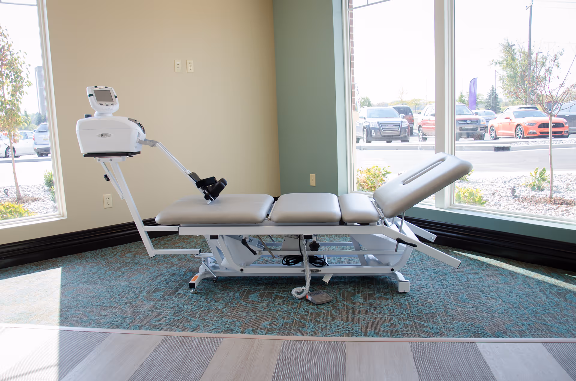A physical therapy treatment table with adjustable sections and attached electronic equipment, placed near large windows showing a parking lot outside.