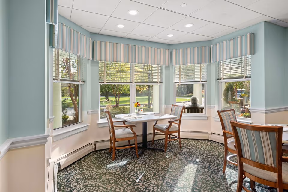 A bright dining area with large windows featuring striped valances, allowing natural light to fill the space. There are wooden chairs with striped cushions around a small square table set with placemats and a small vase of flowers. Outside the windows, greenery and trees are visible.