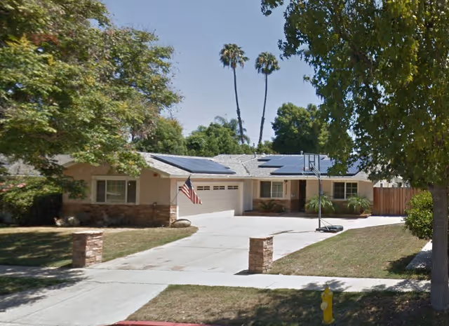 Single-story residential building with a driveway, solar panels, flag, and basketball hoop.