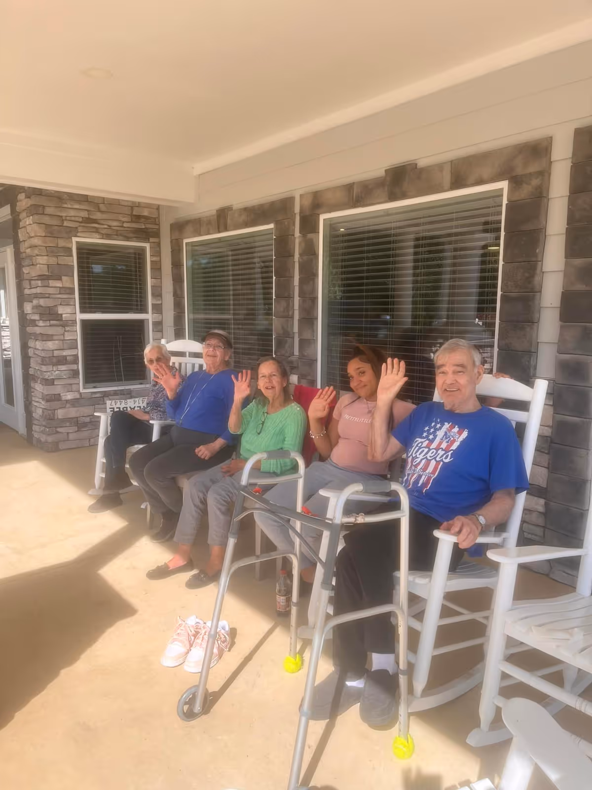 Five elderly individuals sitting on white rocking chairs on a covered porch outside a building with stone and siding walls. They are smiling and waving at the camera. A walker and a pair of pink shoes are visible on the ground in front of them.