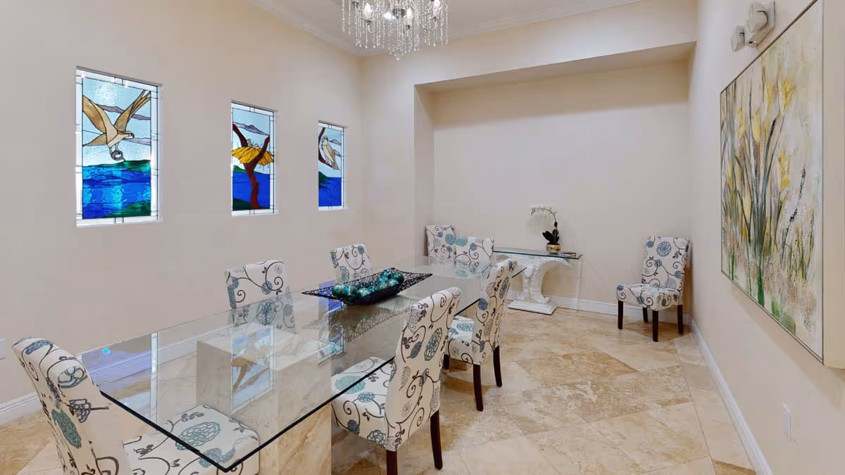 A dining room with a long glass table surrounded by eight upholstered chairs with floral patterns. Three stained glass windows depicting birds and nature are on the left wall. A chandelier hangs from the ceiling, and a large painting of flowers is on the right wall. The floor is tiled with beige stone.
