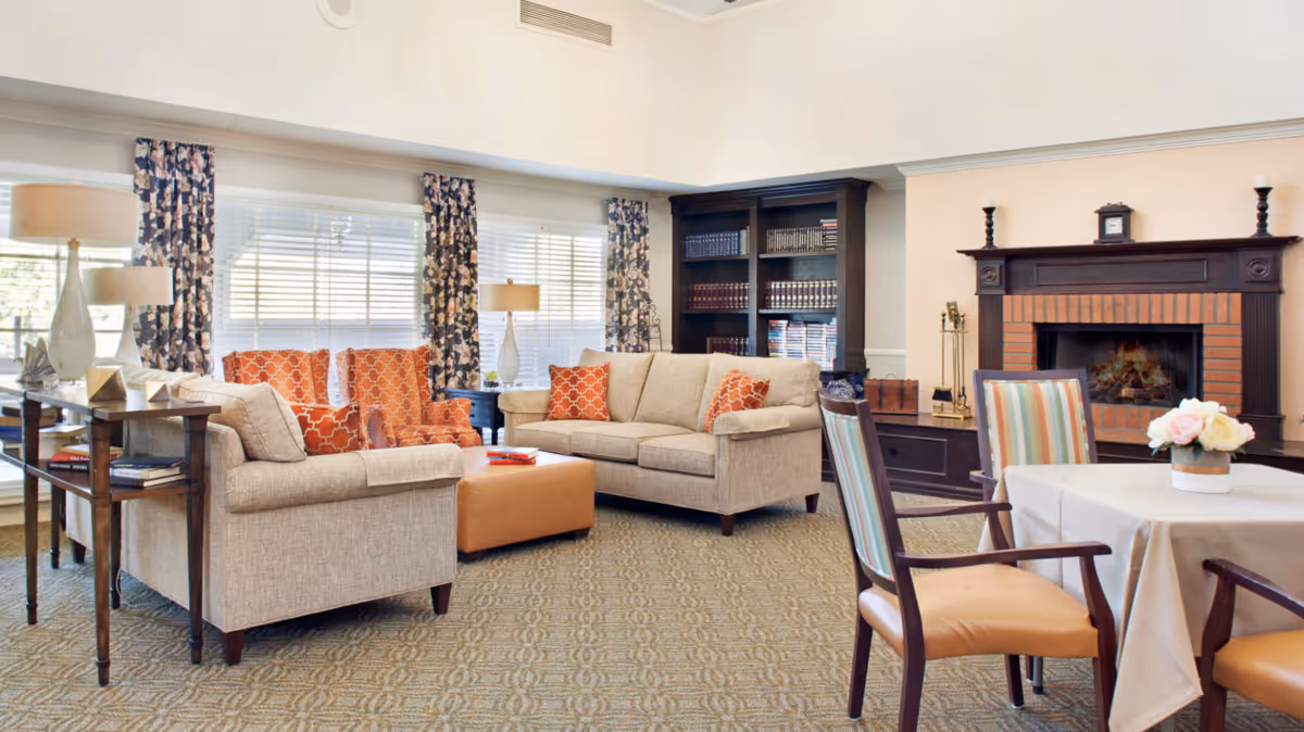 A bright, well-furnished living room in a senior facility with sofas and orange-patterned chairs around a central ottoman, a fireplace, bookshelf and a small dining table.