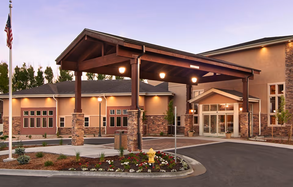 Exterior view of a senior living facility named Gardens on Quail at dusk, featuring a covered entrance with wooden beams and stone pillars, a landscaped area with flowers and a fire hydrant, and an American flag on a flagpole.