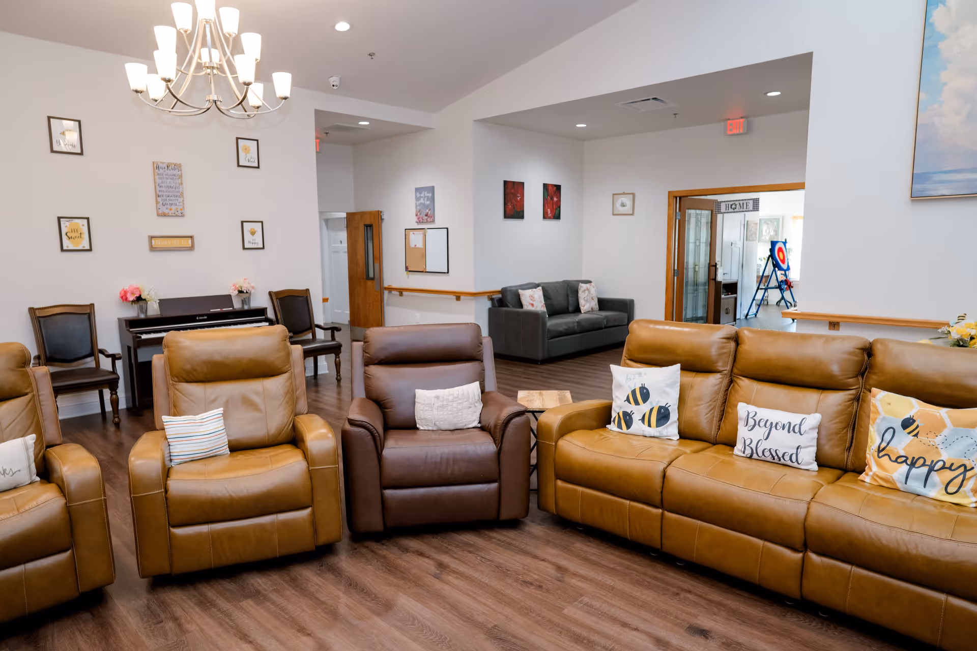 A cozy living room area in a senior living facility with brown leather recliners and a matching sofa adorned with decorative pillows. The room features wood flooring, a chandelier, framed wall art, a piano with chairs, and a gray couch in the background. There is a doorway leading to another room with a target board and a sign that says 'HOME'.
