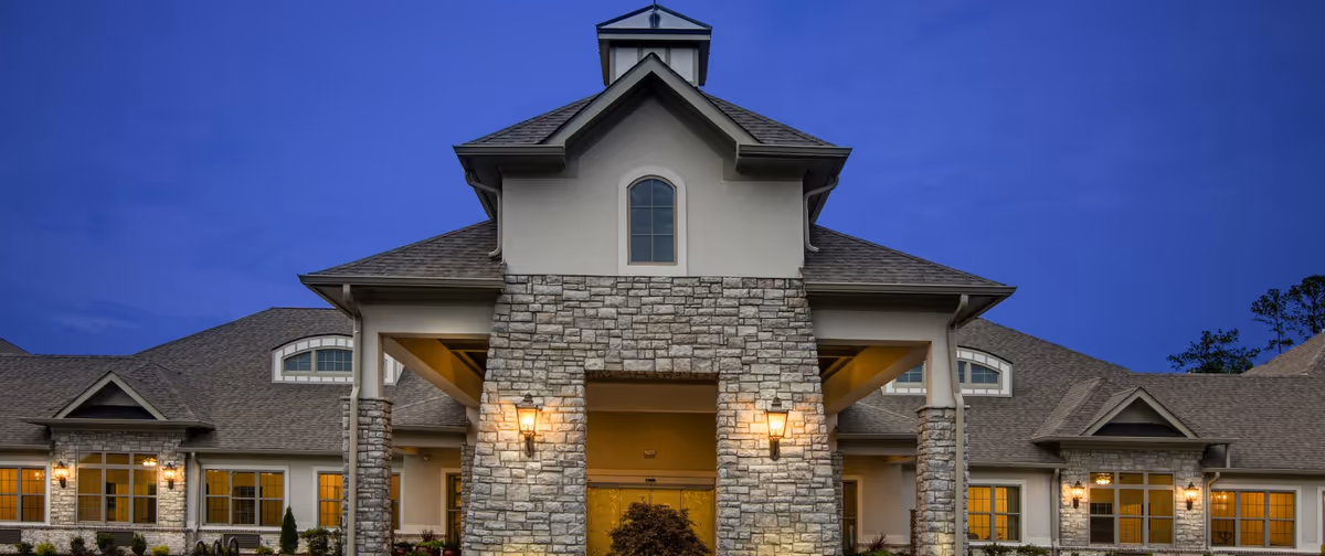 Front exterior view of a senior living facility building named Orchard at Athens during twilight, featuring stone and beige walls, multiple windows with warm interior lighting, and a peaked roof with a small cupola.