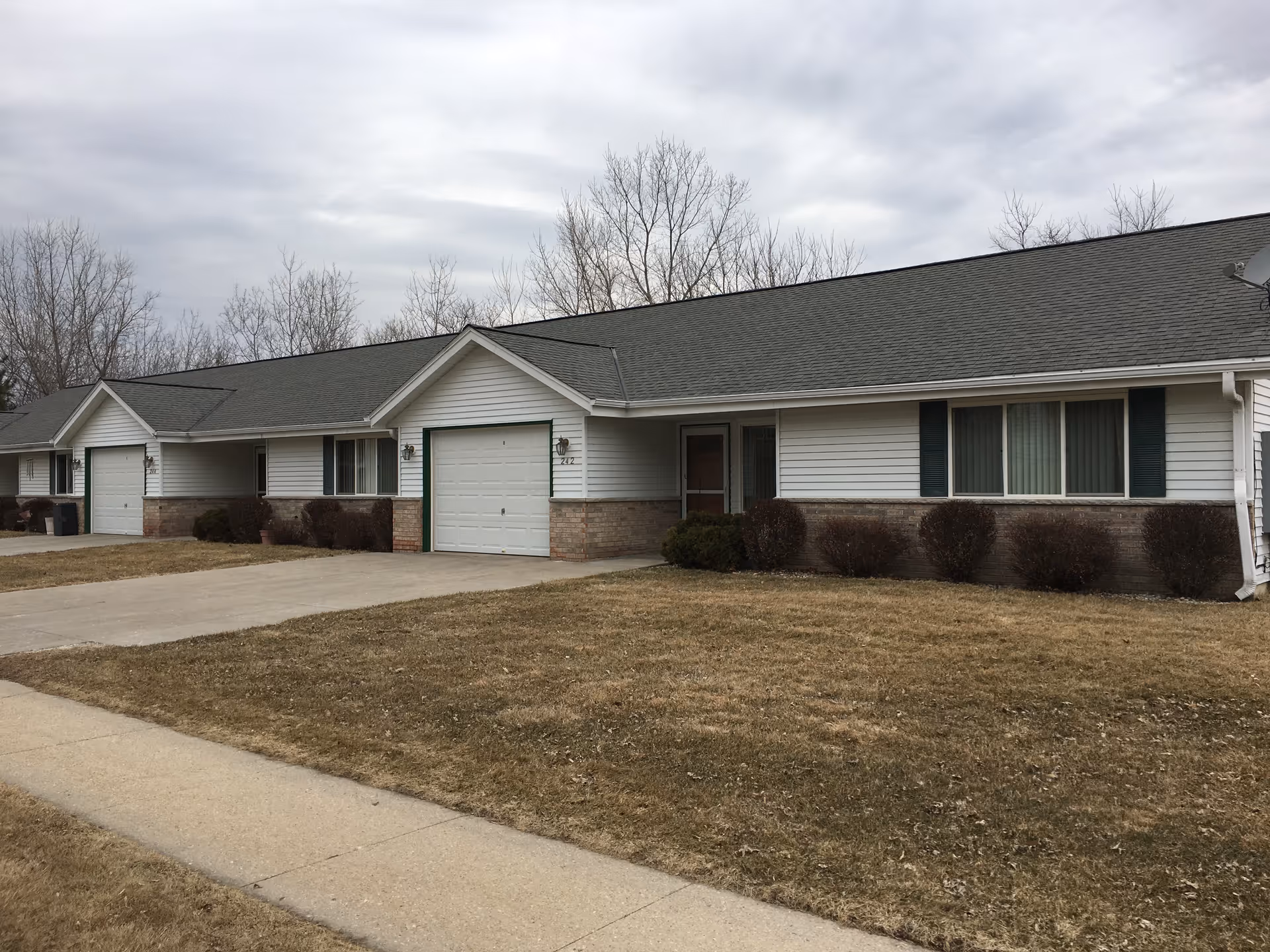 Exterior view of a single-story residential building with white siding, green shutters, and attached garages. The building is surrounded by a dry lawn and leafless trees under a cloudy sky.