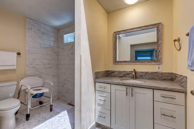 Accessible bathroom with a walk-in tiled shower and shower chair, toilet, and a vanity with sink and mirror.