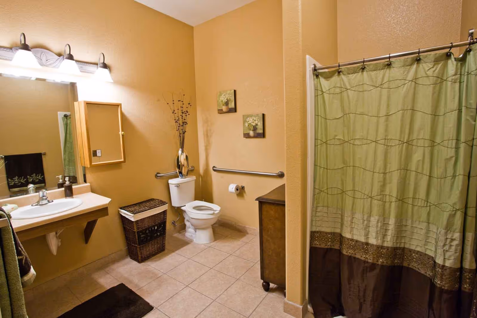 A bathroom with beige walls and tiled floor featuring a white sink with a mirror and light fixture above it on the left, a wicker laundry basket next to a white toilet with a grab bar behind it, two small framed floral pictures on the wall, a dark brown cabinet on wheels, and a shower with a green and brown patterned curtain on the right.