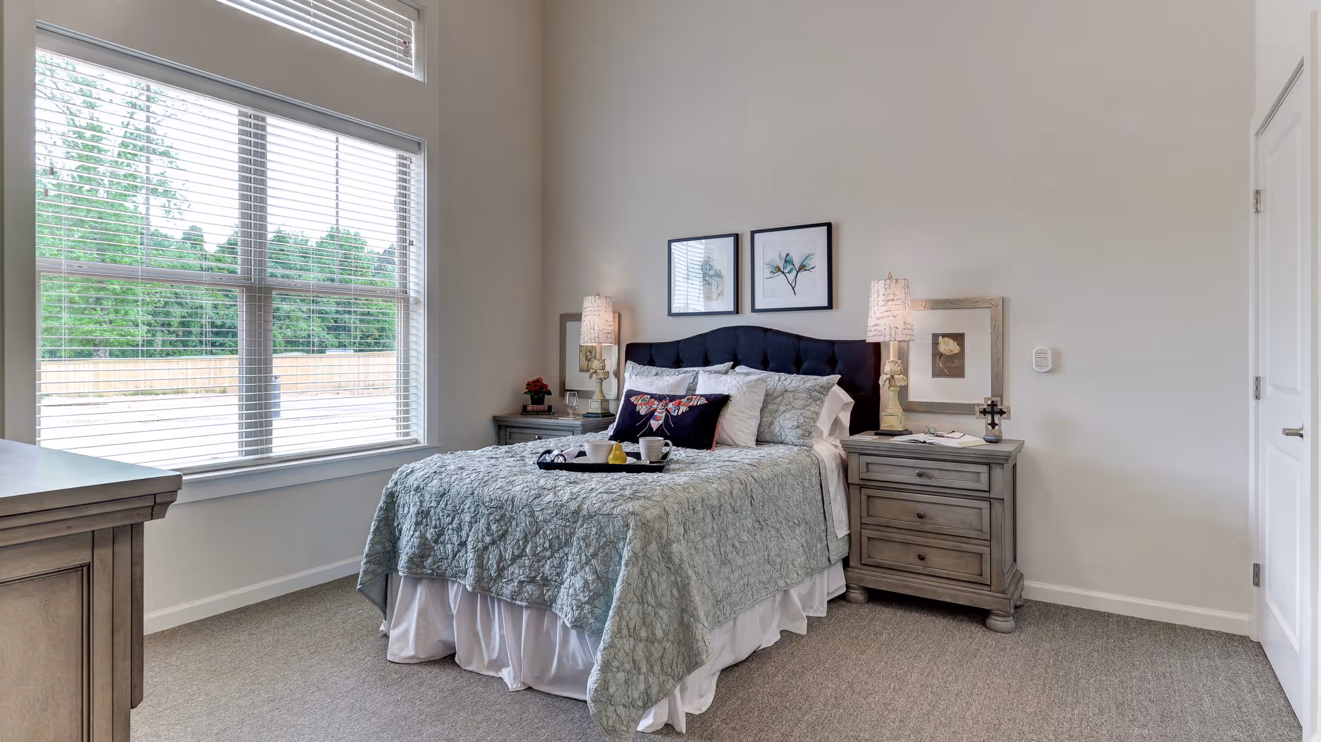 Well-lit bedroom with a made bed, tufted headboard, two nightstands with lamps, and a large window overlooking trees.
