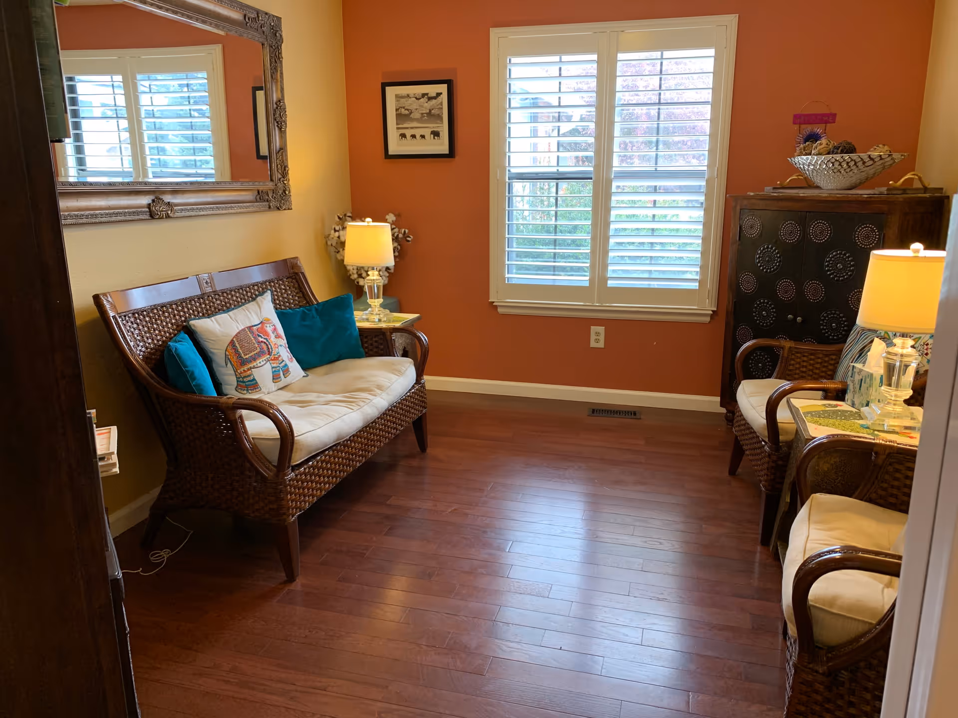 A cozy sitting area with wooden flooring, featuring a wicker loveseat and two wicker armchairs with cushions. There are two table lamps on side tables, a large decorative mirror on one wall, a framed picture, and a window with white shutters letting in natural light. A decorative cabinet with a bowl of pinecones is also visible.