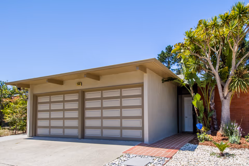 Exterior view of a single-story building with a double garage door, a tiled walkway leading to a white entrance door, and landscaping with trees and plants under a clear blue sky.