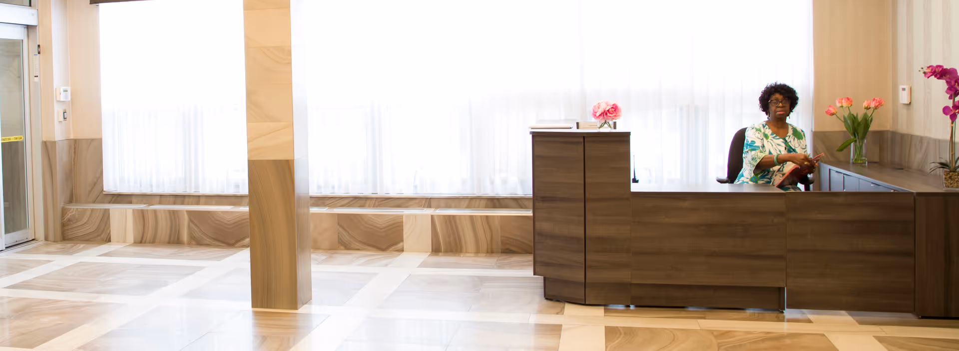 Bright lobby interior with a reception desk and a seated receptionist by large curtained windows.