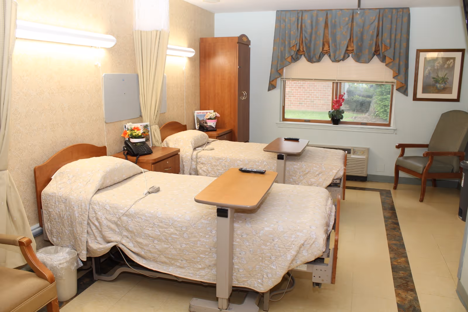 Two hospital-style beds in a shared senior care room with overbed tables, a window, and chairs.