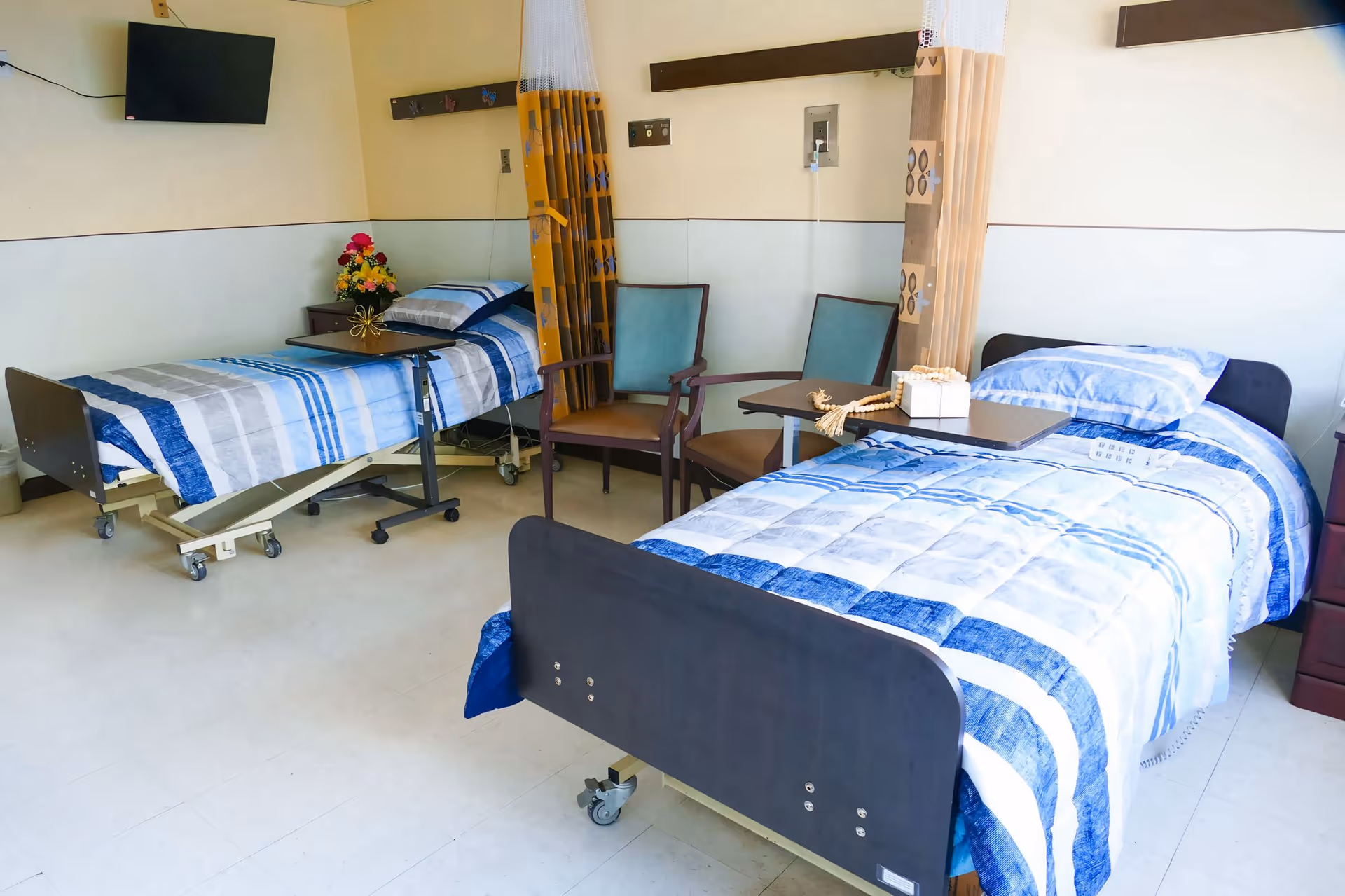 A room with two hospital-style beds, each with blue and white striped bedding. Between the beds are two chairs and a small table with a decorative item on it. There is a wall-mounted TV in the corner and a curtain divider between the beds.
