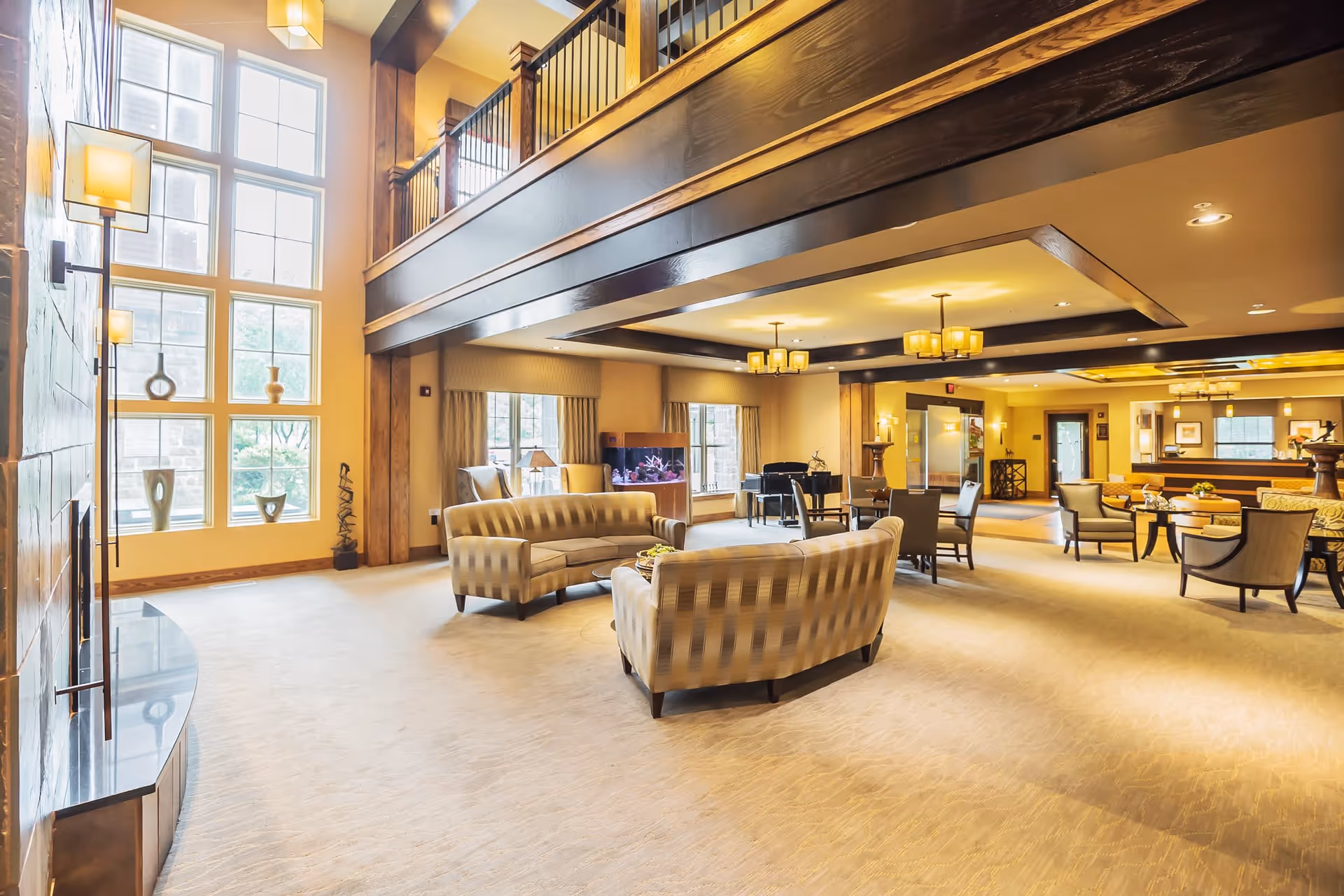 Spacious senior living facility common area with high ceilings and large windows allowing natural light. The room features multiple seating arrangements including sofas and chairs, a fireplace, decorative vases on window ledges, and warm lighting fixtures. There is a piano in the background and a second-floor balcony overlooking the area.