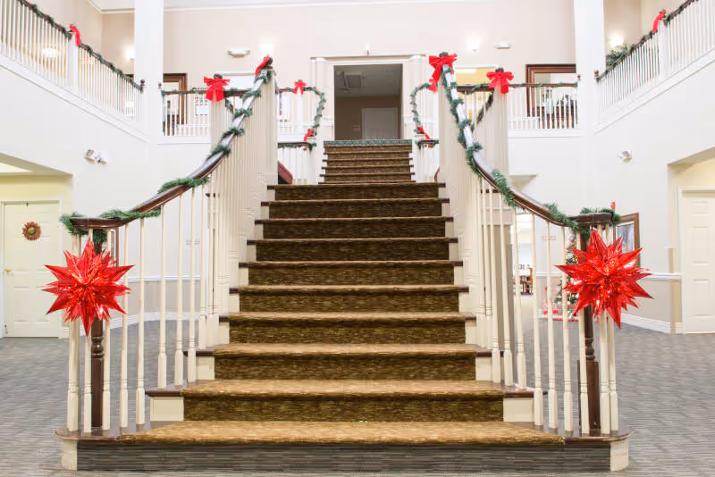 Wide staircase in a senior living facility decorated with green garlands and red bows, with large red star ornaments on the banisters. The staircase leads to an upper floor with white railings and beige walls.